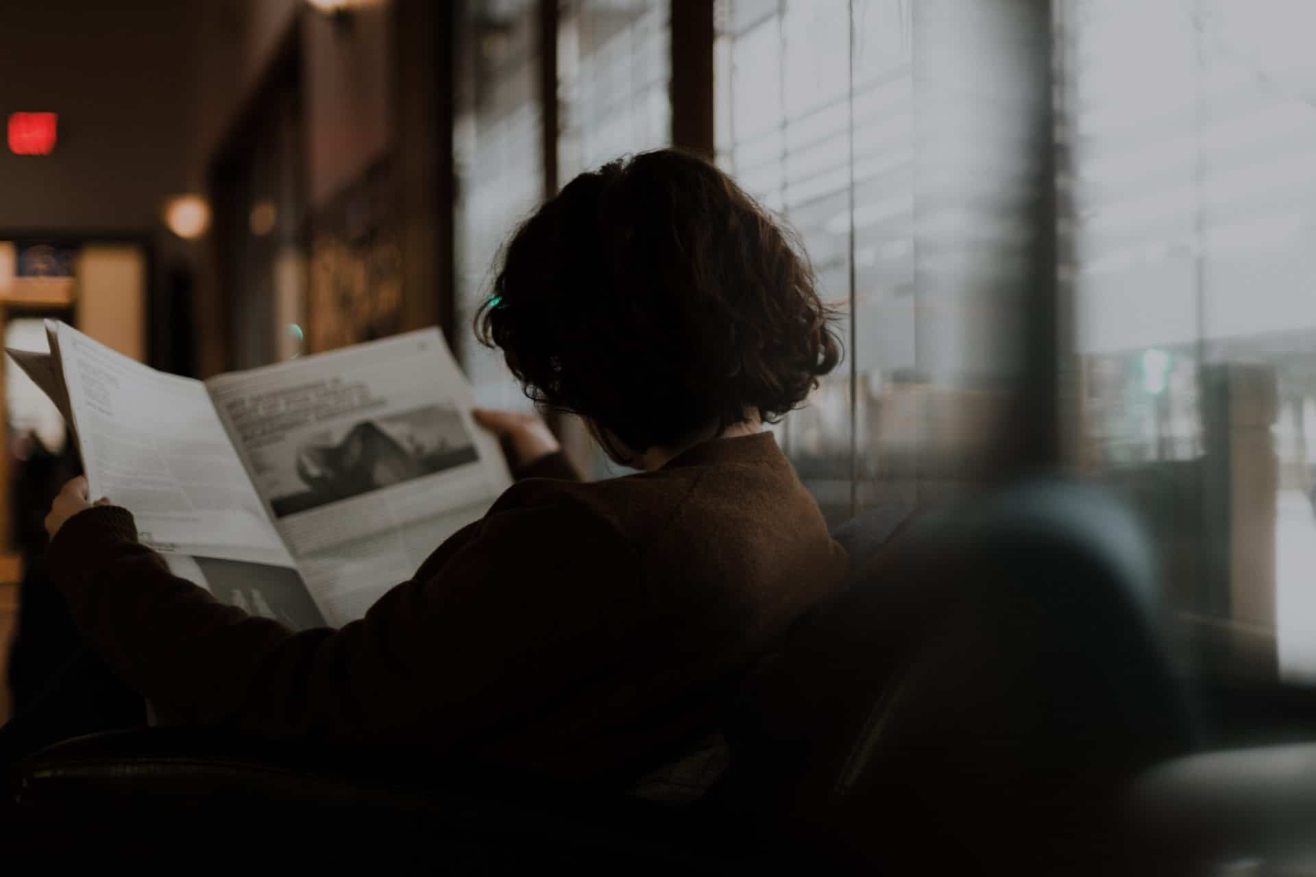 woman sitting on couch and reading the newspaper with her back facing us.