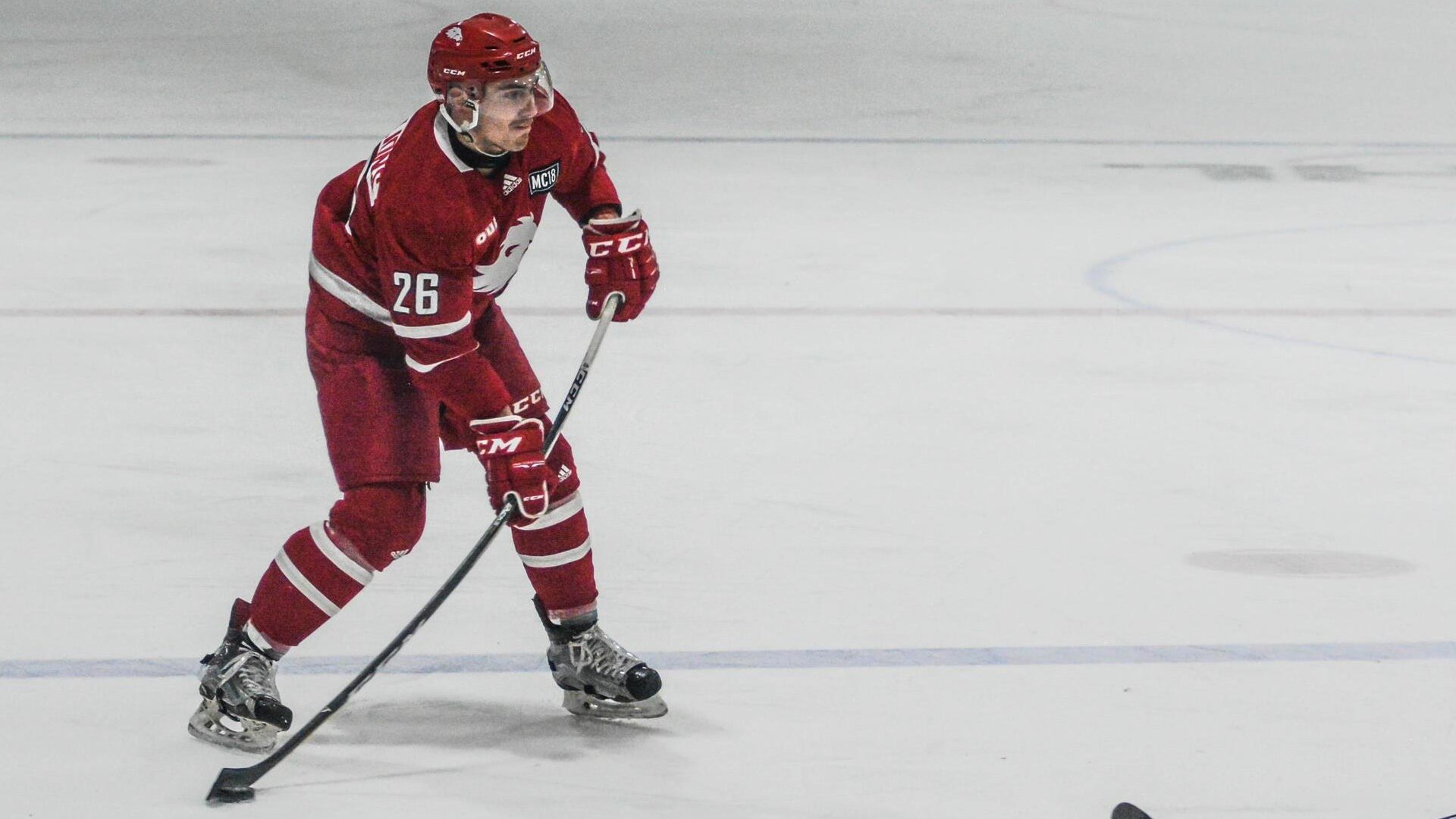 A man in a red jersey holds a puck on his stick while playing on ice with skates.
