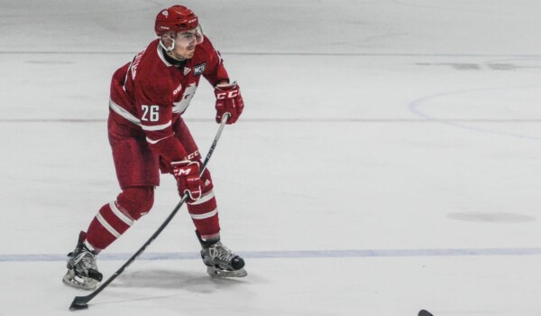 A man in a red jersey holds a puck on his stick while playing on ice with skates.