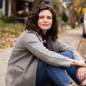 Maighdlin has brown hair and blue eyes, and is sitting outside in jeans and a grey coat